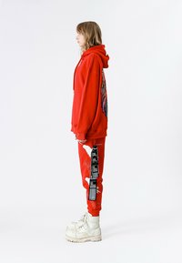 Sudadera con capucha roja con diseño de alas multicolores en la espalda, combinada con pantalones rojos con detalles blancos y una etiqueta. Se lleva con botas blancas gruesas.