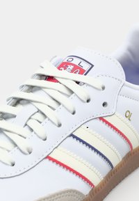 Baskets blanches avec des rayures bleues, rouges et crème, présentant un patch logo et une semelle en caoutchouc texturé de couleur marron clair. Les lacets sont blancs.