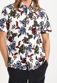 Camicia a maniche corte con sfondo bianco e fantasie floreali colorate in blu, rosso e giallo. Design con bottoni e vestibilità comoda.