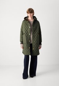 Gequilted groene parka met een capuchon, voorzien van gouden accenten, zijzakken, en een gestructureerde gebreide trui eronder, gecombineerd met uitlopende donkere denim.