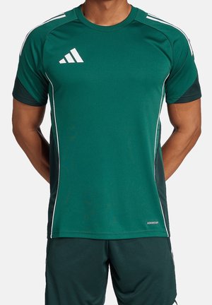 Homme portant un maillot de sport vert Adidas et un short assorti avec des bandes blanches, debout les bras derrière le dos.