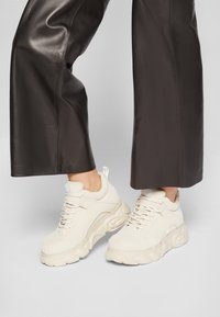 Beiga sneakers med en texturerad läderöverdel, snörningsdesign och en kraftig gummisula med ett vågformat mönster.