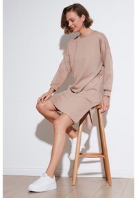 Knee-length, beige sweatshirt dress with long sleeves and side slits, worn with white sneakers while seated on a wooden stool.