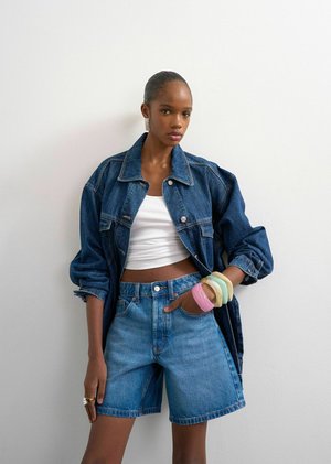 Jeune femme debout contre un mur blanc, portant une veste en jean oversize, un short en jean taille haute, un top court blanc et des bracelets colorés à un poignet.