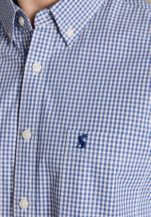 Mężczyzna ubrany w niebiesko-białą, drobną kratkę koszuli na guziki z kołnierzykiem i przednią kieszenią z granatowym haftowanym logo zwierzęcia.