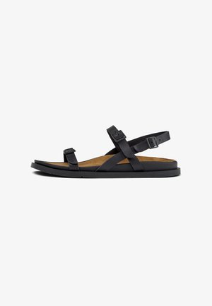 Black leather sandal featuring crossed straps, adjustable buckles, a cushioned insole, and a flat rubber sole.