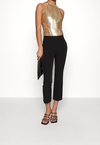 Woman wearing a gold sequined sleeveless top, black cropped pants, black strappy heels, holding a black clutch against a white background.