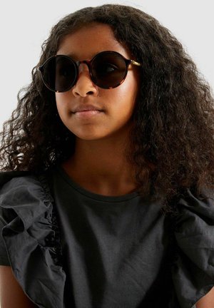 Lunettes de soleil écaille avec verres ronds, monture noire et accents dorés ; portées avec une chemise foncée à manches volantées.