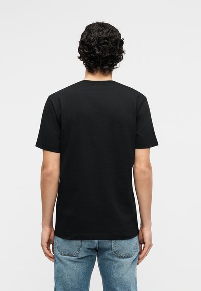 Άνδρας με σγουρά σκούρα μαλλιά, ντυμένος με απλό μαύρο κοντομάνικο μπλουζάκι και μπλε τζιν, στέκεται με την πλάτη γυρισμένη προς ένα απλό φόντο.