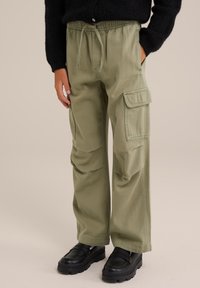 Olīvu zaļas kravas bikses ar brīvu piegriezumu, elastīgu jostu, auklu un vairākām sānu kabatām, kombinētas ar melnām plakanām kurpēm.