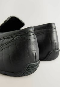 Zwarte leren schoenen met een gestructureerde afwerking, met een laag rugontwerp en rubberen zool voor grip. Stikseldetails zichtbaar langs de randen.