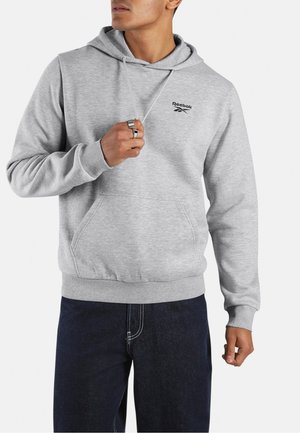 Man wearing a light gray Reebok hoodie with a front pocket and dark blue jeans, holding the hoodie string near the neck.
