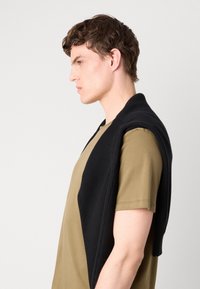 Svart strikket vest drapert over en khaki t-skjorte, med ribbestruktur og åpen design, vist mot en nøytral bakgrunn.