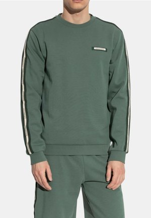 Uomo che indossa una felpa verde a maniche lunghe e pantaloni abbinati con dettagli a strisce bianche e nere sulle maniche e sui lati dei pantaloni.