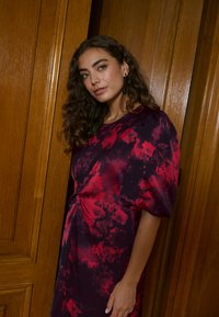 Fialové a červené květované šaty s volným střihem a nafouklými rukávy, zdobené překrouceným detailem vpředu. Hladká, lehká látka.