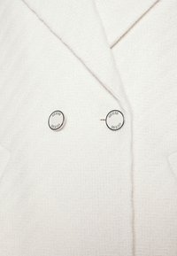 Blazer blanco texturizado con patrón de espiga. Dos botones redondos con el logo "GUESS" en detalles en blanco y negro cerca del cuello.