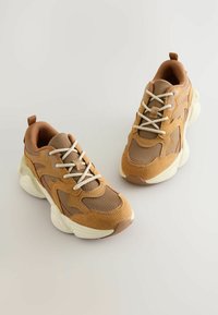 Brązowo-tanowe sneakersy wykonane z siateczki i zamszu, z białymi sznurówkami i masywną, teksturowaną podeszwą. Minimalistyczny design, zaokrąglony kształt.