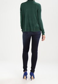 Pull vert à coupe ample avec un col montant, associé à un jean skinny noir et des chaussures à talons bleues. Tissu lisse et lignes épurées.