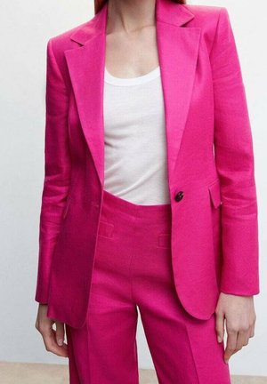 Kvinde iført en knaldrød pink blaz jakke og matchende bukser over en hvid ribbet tanktop, stående mod en ensfarvet lys baggrund.