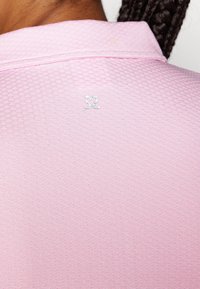 Camisa polo rosa clara com tecido texturizado, apresentando um pequeno logótipo de coração prateado e ossos cruzados na parte de trás da gola.