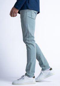 Pantalones verde claro, ajustados, con costuras sutiles y bolsillos traseros, combinados con zapatillas blancas de suela texturada, sobre un fondo liso.