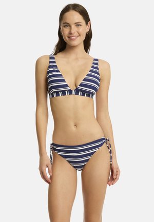 Femme aux longs cheveux bruns portant un bikini rayé bleu marine et blanc avec des côtés noués, debout et souriante sur un fond clair uni.