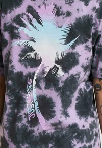 Tricou tie-dye în nuanțe de violet și negru, având un grafic cu o palmier în bleu deschis și textul "Just Rise" în culori pastelate.