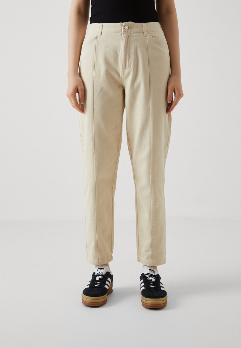 Beige, högmidjade byxor med rak passform, utrustade med sidosisar och fickor, i kombination med svarta sneakers och vita strumpor.