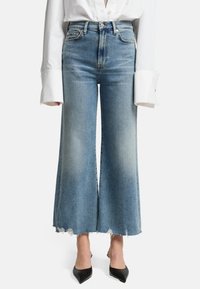 Jeans a vita alta di colore blu chiaro, svasati con orlo grezzo e leggera scoloritura; abbinati a una camicia bianca oversize e a delle ballerine nere a punta.
