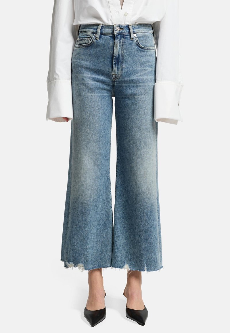 Jeans a vita alta di colore blu chiaro, svasati con orlo grezzo e leggera scoloritura; abbinati a una camicia bianca oversize e a delle ballerine nere a punta.