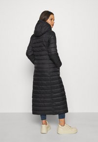 Long manteau d'hiver noir avec une capuche, design matelassé et texture côtelée. Porté avec un jean bleu et des baskets à plateforme beiges.