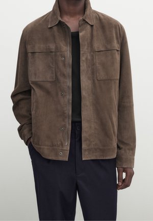 Man draagt een bruine suède jas met voorzakken over een zwart shirt en donkere broek, staand tegen een effen achtergrond.