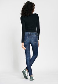 Skinny jeans in dark wash denim, featuring a mid-rise waist, two back pockets with stitching detail, paired with white and green sneakers.