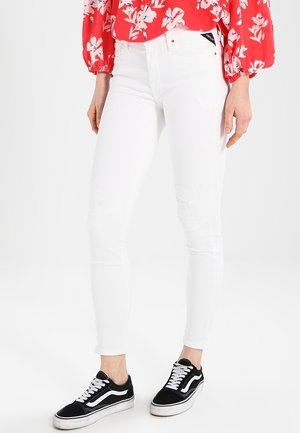Persoon die witte skinny jeans, zwart-witte sneakers en een rode bloemenlange mouwen blouse draagt, staand tegen een witte achtergrond.