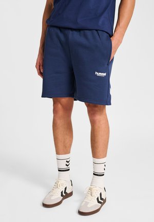 Mann in marineblauen Hummel-Shorts, weißen Socken mit schwarzen Streifen und weißen Sneakern mit schwarzen Akzenten, steht mit einer Hand in der Tasche.