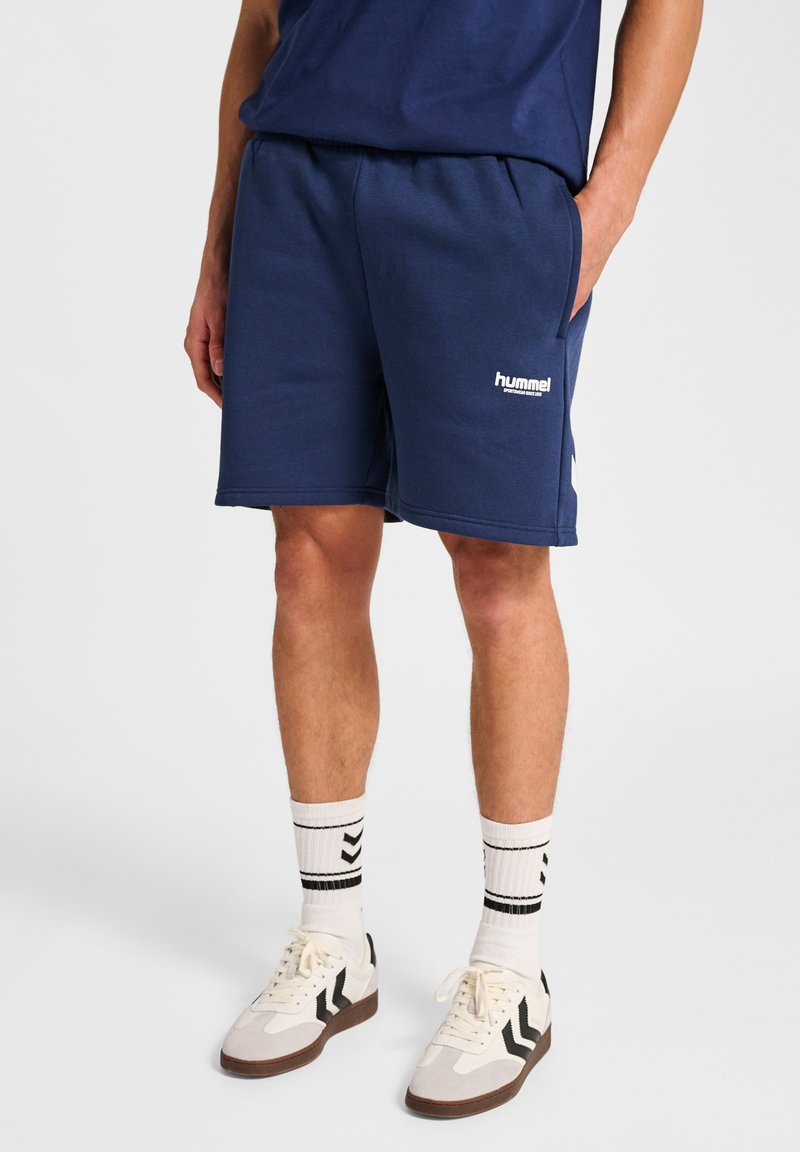 Man som bär marinblå Hummel-shorts, vita strumpor med svarta ränder och vita sneakers med svarta detaljer, som står med en hand i fickan.