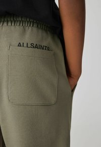 Olivno zeleni trenerki z elastičnim pasom, ki imata zadnji žep in natisnjen logotip "ALLSAINTS." v črni barvi nad žepom.