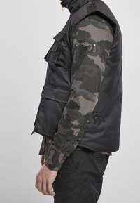 Černá vesta s kapsami, vrstvená přes dlouhý rukáv s maskáčovým vzorem. Má knoflíky a texturovaný povrch látky.