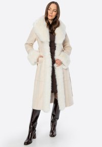 Beige wollen jas met een witte faux fur kraag en manchetten. Lang, getailleerd ontwerp met een strik taille en een gladde textuur. Bruine laarzen tot de knie.