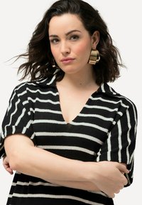 Haut rayé noir et blanc avec un col en V, des manches courtes et un tissu texturé. Le modèle porte de grandes boucles d'oreilles en forme de créole en or.
