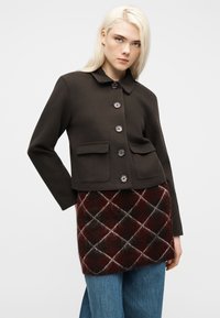 Νεαρός με ξανθά μαλλιά φοράει σκούρο καφέ σακάκι με κουμπιά, καρώ φούστα και μπλε τζιν, στέκεται μπροστά σε λευκό απλό φόντο.