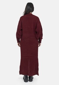 Cardigan lungo borgogna lavorato a maglia con un motivo testurizzato, maniche larghe e taglio dritto, indossato sopra stivali neri. Nessuna chiusura visibile.