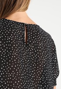 Černá průhledná halenka s bílým skvrnitým vzorem, se zapínáním ve tvaru klíčové dírky na zádech a jemným, volně splývajícím materiálem.