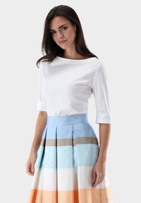 Weißes, langärmeliges Oberteil kombiniert mit einem plissierten, mehrfarbigen Rock mit hellblauen, beigen, weißen und orangen horizontalen Streifen.