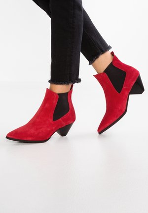 Osoba ubrana w czarne jeansy z postrzępionymi brzegami oraz czerwone zamszowe botki na koturnie z czarnymi elastycznymi wstawkami po bokach i niskim, masywnym obcasie.