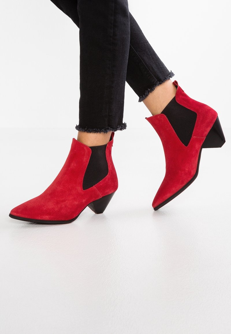 Personne portant un jean noir à ourlet effiloché et des bottines en daim rouge à bout pointu avec des panneaux latéraux élastiques noirs et des talons bas épais.