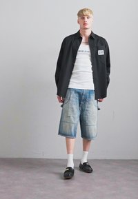 Ung man som står med en svart öppen skjorta, vit linne, långa denimshorts, vita strumpor och svarta loafers mot en slät vägg.