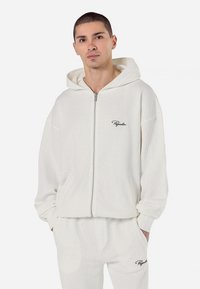 White corduroy zip-up hoodie with a hood, ribbed texture, and a small, dark logo on the chest. Relaxed fit and long sleeves.
