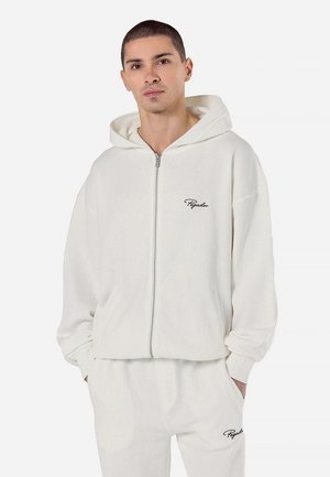 White corduroy zip-up hoodie with a hood, ribbed texture, and a small, dark logo on the chest. Relaxed fit and long sleeves.
