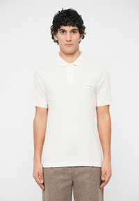 Νεαρός άντρας με σγουρά μαλλιά, φορώντας ένα απλό λευκό polo shirt με ετικέτα "Armani Exchange" και ανοιχτόχρωμο καφέ υφασμάτινο παντελόνι.